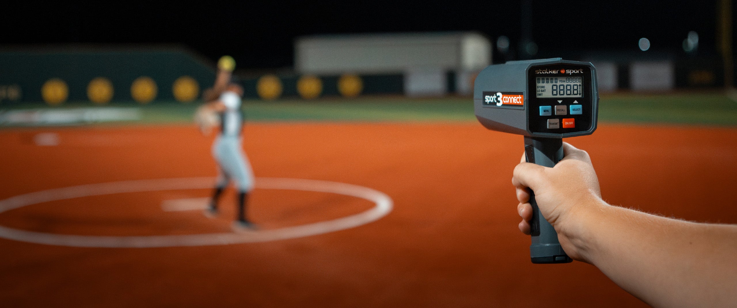 A person holding a Sport 3 Connect radar measuring the velocity of a pitch being thrown by a softball pitcher in the background.