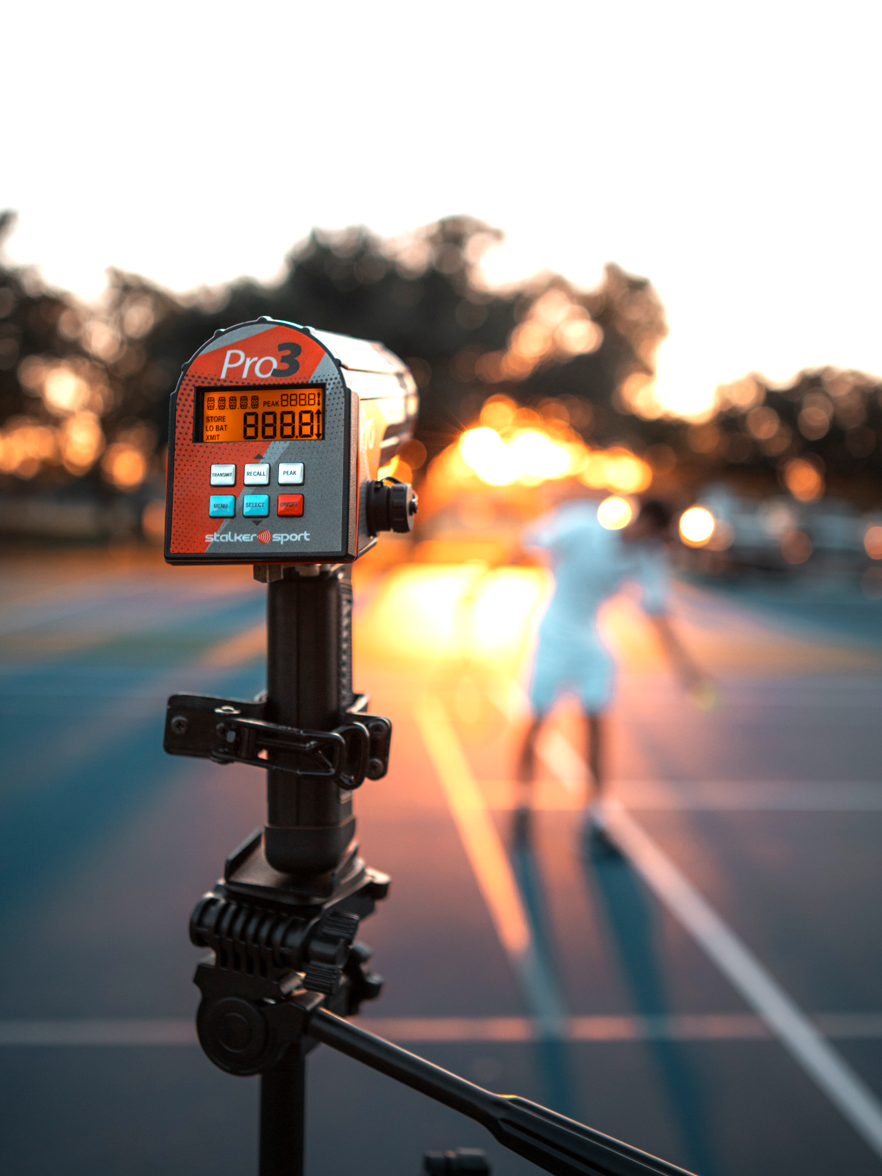A Stalker Sport Pro 3 radar gun mounted to a tripod with a tennis player getting ready to serve in the background.