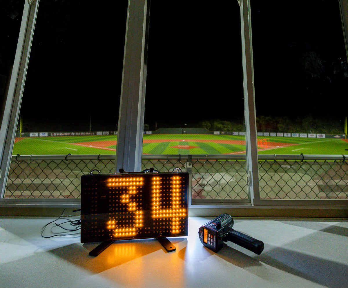 A Stalker Sport Pro 3s radar gun next to a 2.5 digit LED speed sign up in the press box of a baseball field at night.