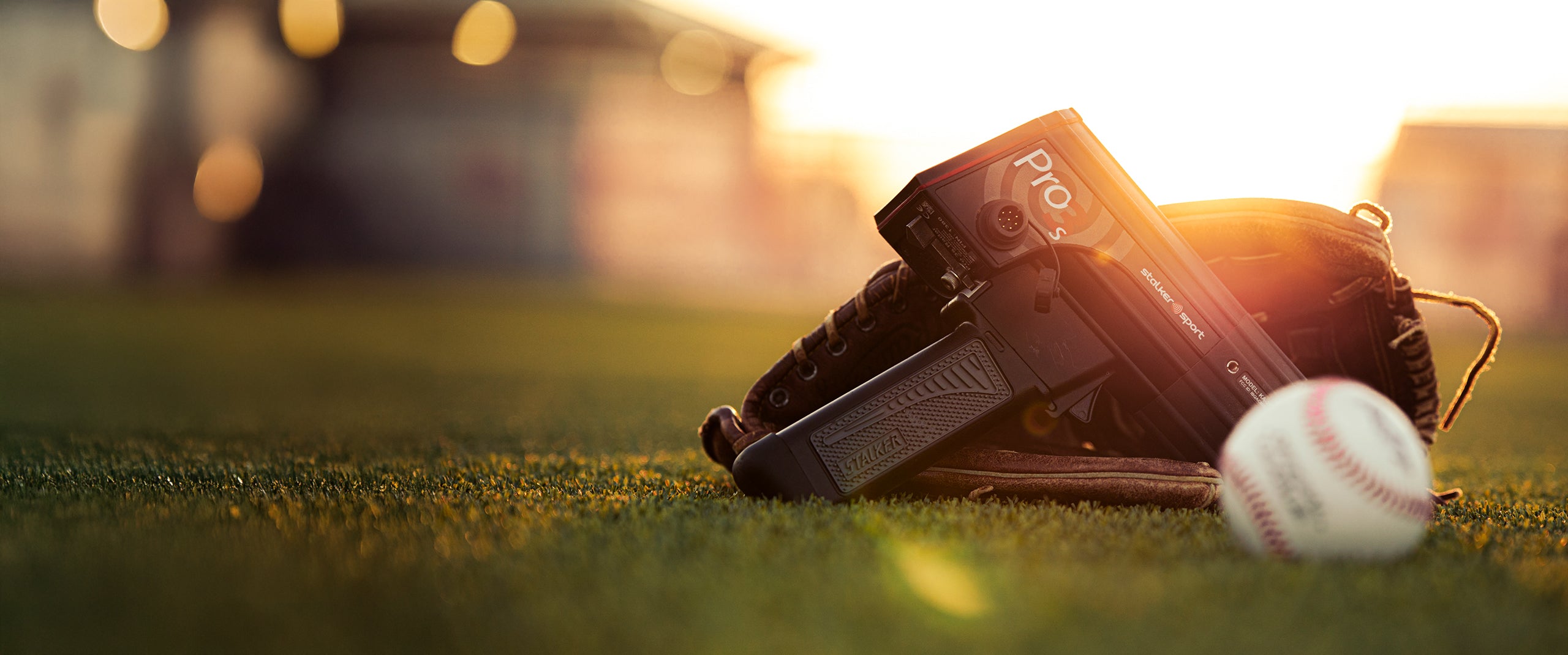 A Pro 3s radar gun with a baseball glove on a baseball field next to a baseball at sunset.