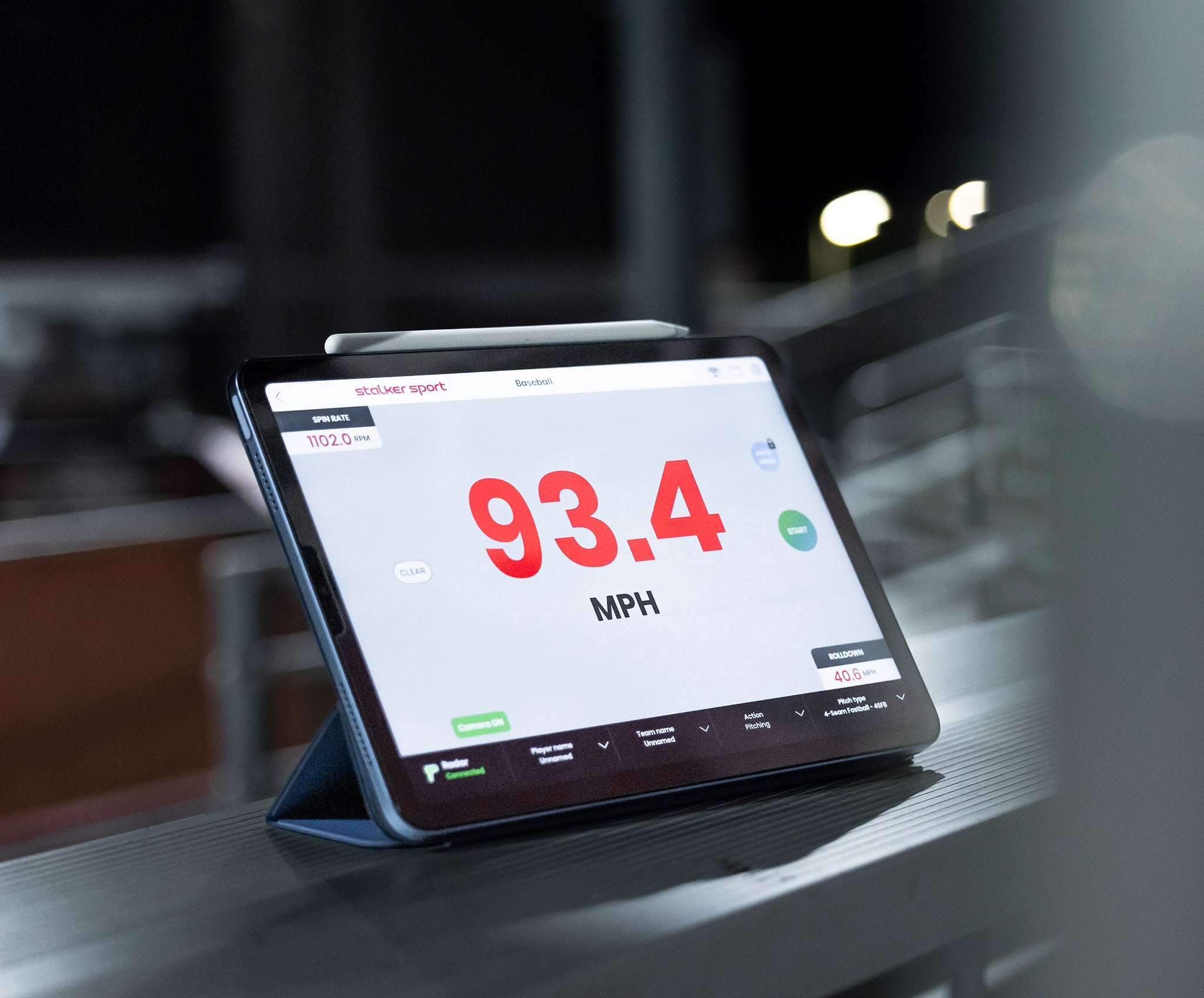 The Stalker Sport Radar app on an iPad resting on the bleachers at a baseball field. The app is displaying a speed of 93.4 MPH with a spin rate of 1102.0 RPM.
