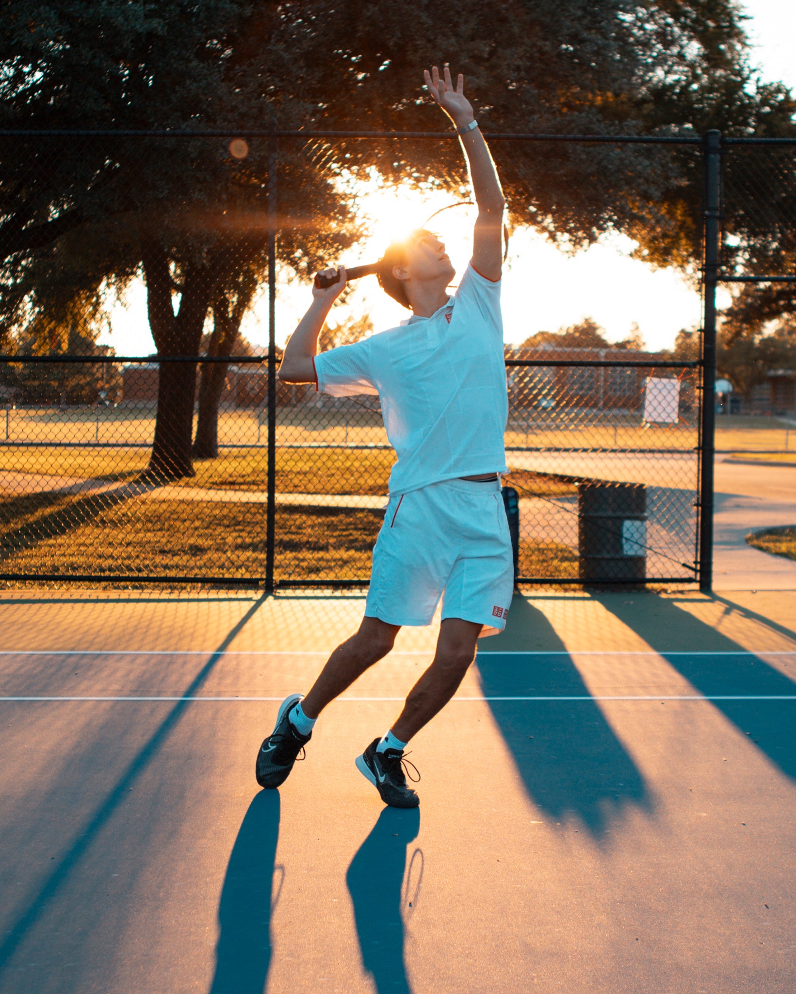 A man from Team Stalker Sport hitting a tennis serve during golden hour with the sun directly behind him.