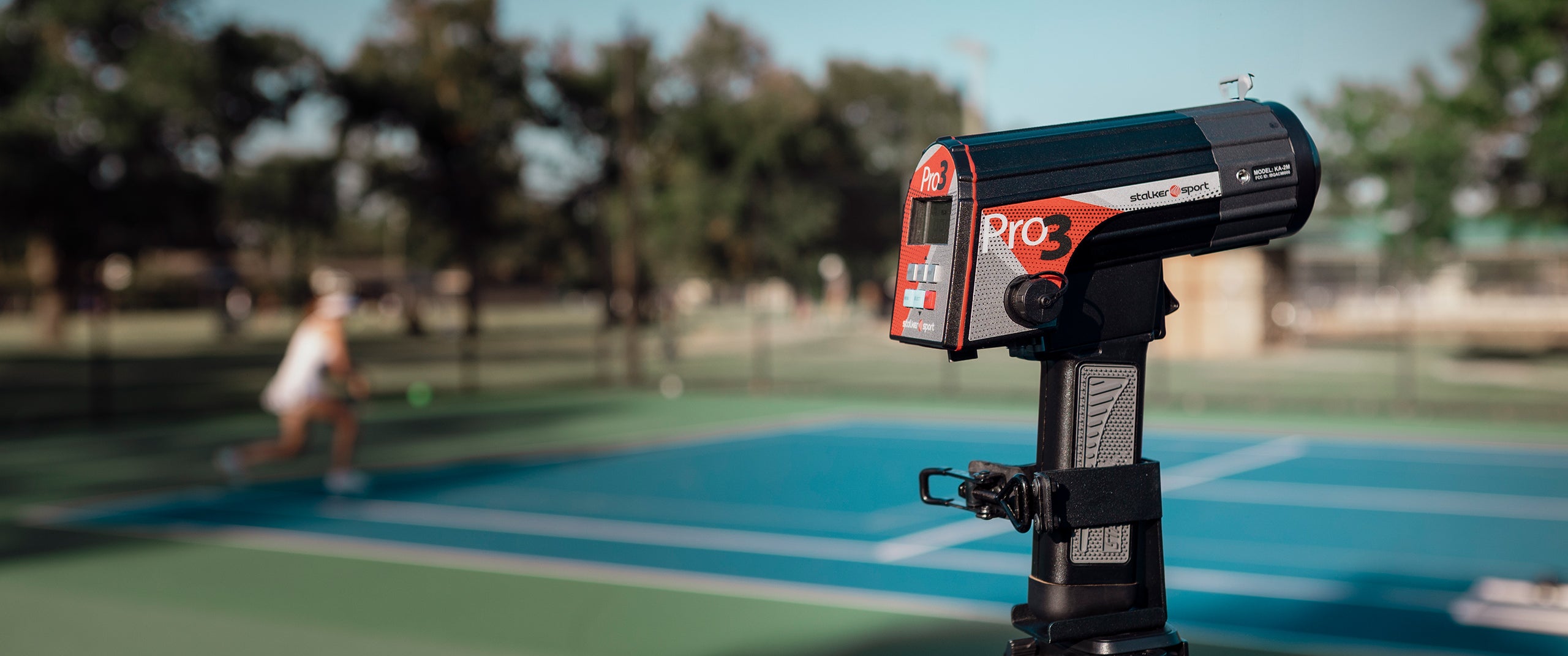 A Pro 3 sport radar gun in a tripod mount in front of a tennis court with a female tennis player in the background.