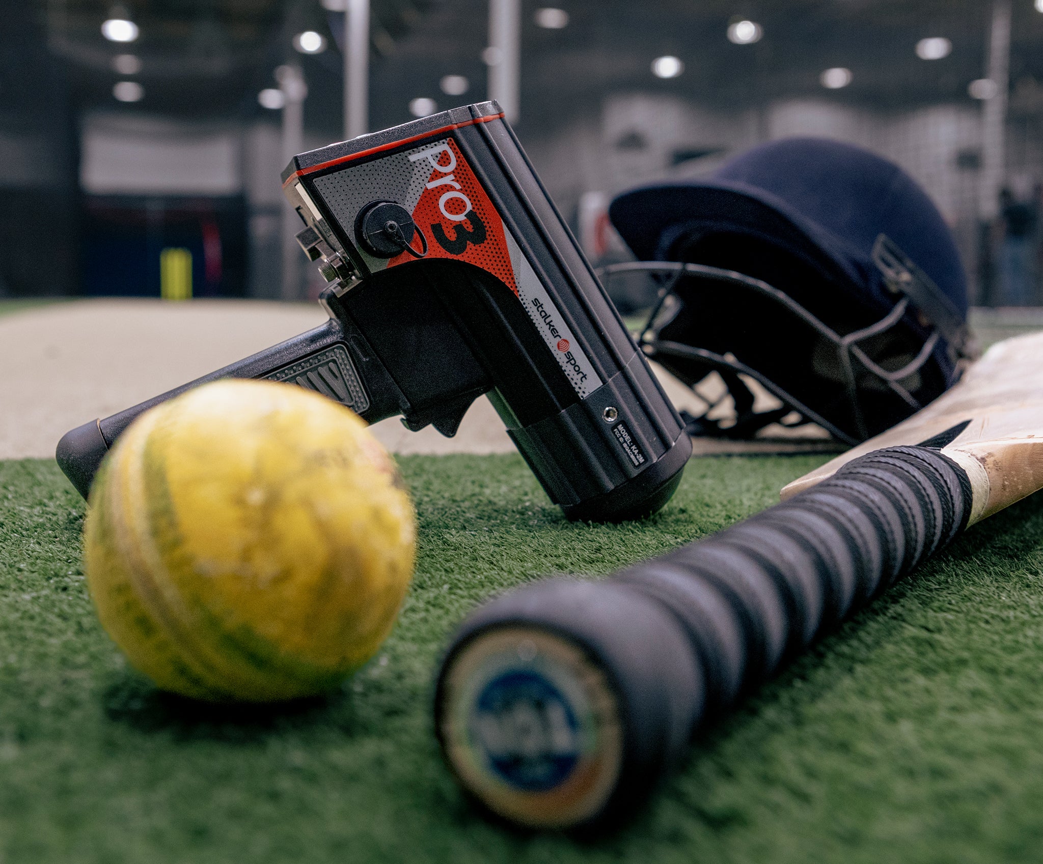 A Stalker Pro 3 radar gun next to a cricket ball, bat, and helmet inside a training facility.