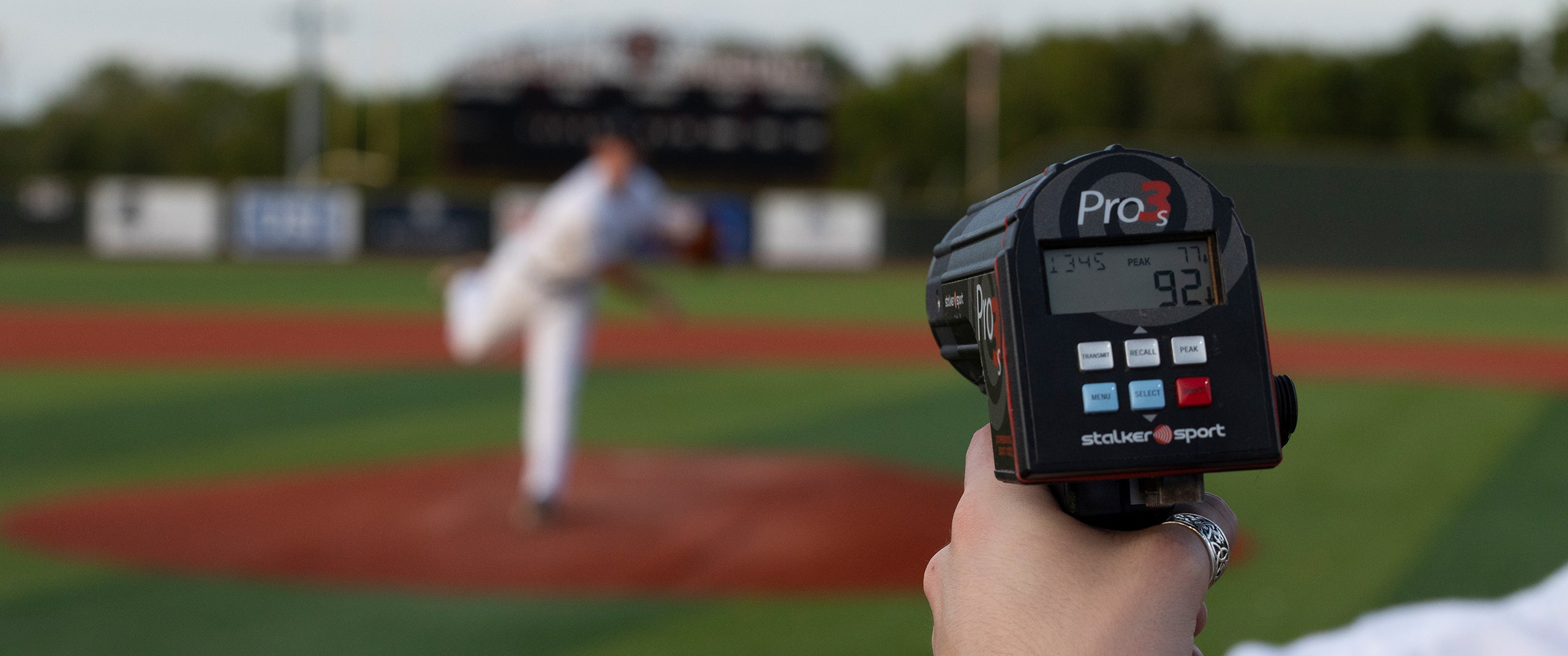 A hand holding a Pro 3s sport radar gun showing a velocity of 92 MPH and a spin rate of 1345 and an over-the-plate speed of 77 MPH. There is a baseball pitcher in the background. 