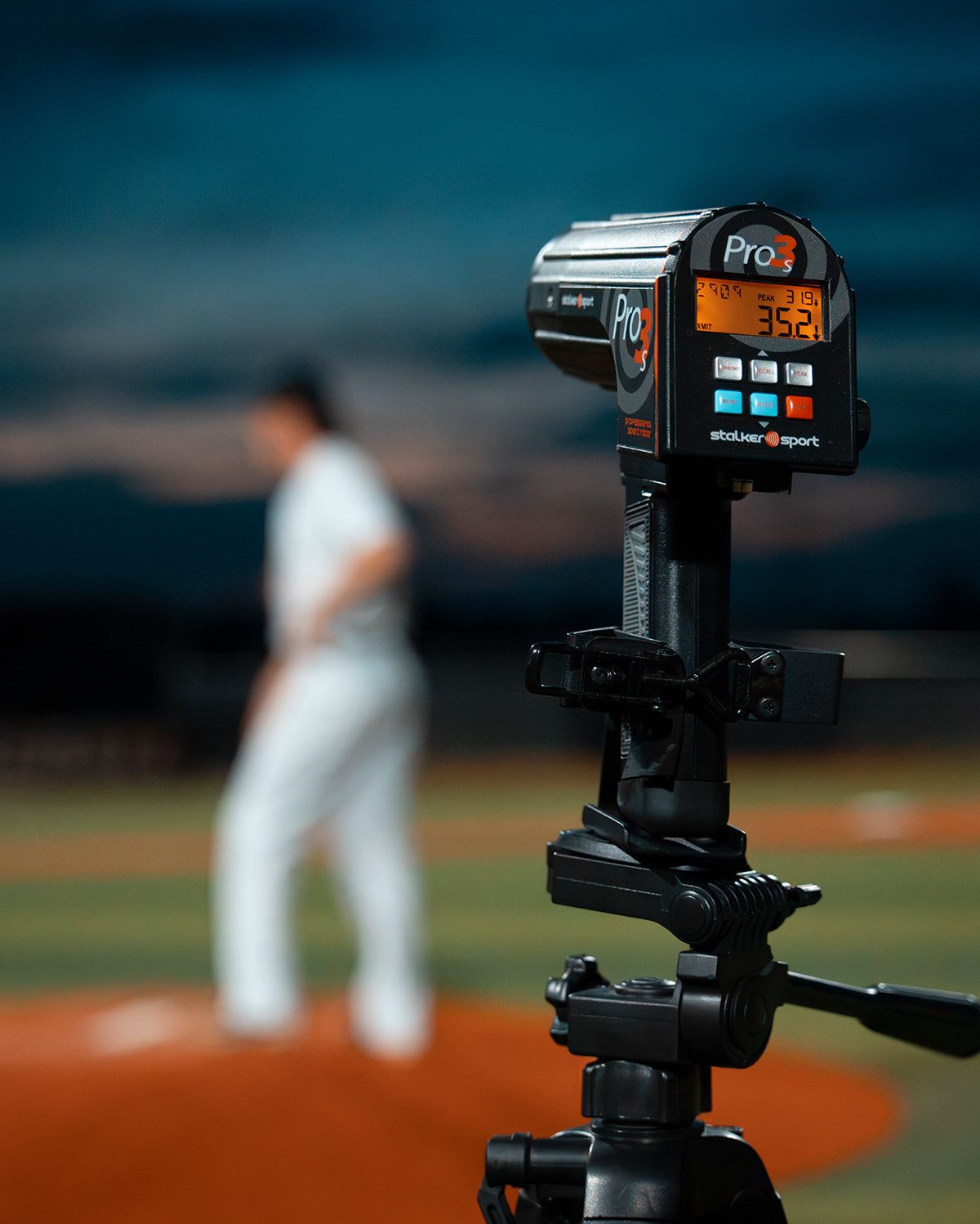 A Pro 3s sport radar gun mounted to a tripod on a baseball field at night.