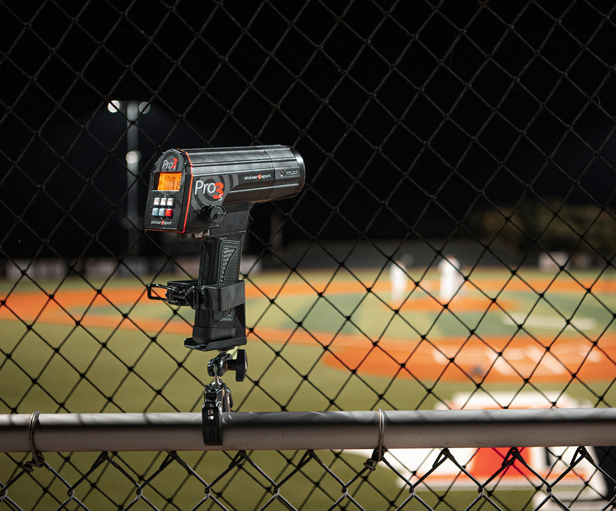 A Pro 3s sports radar gun from Stalker Sport shown in the 4-in-1 mount clamped to a chain link fence with a baseball game in the background.