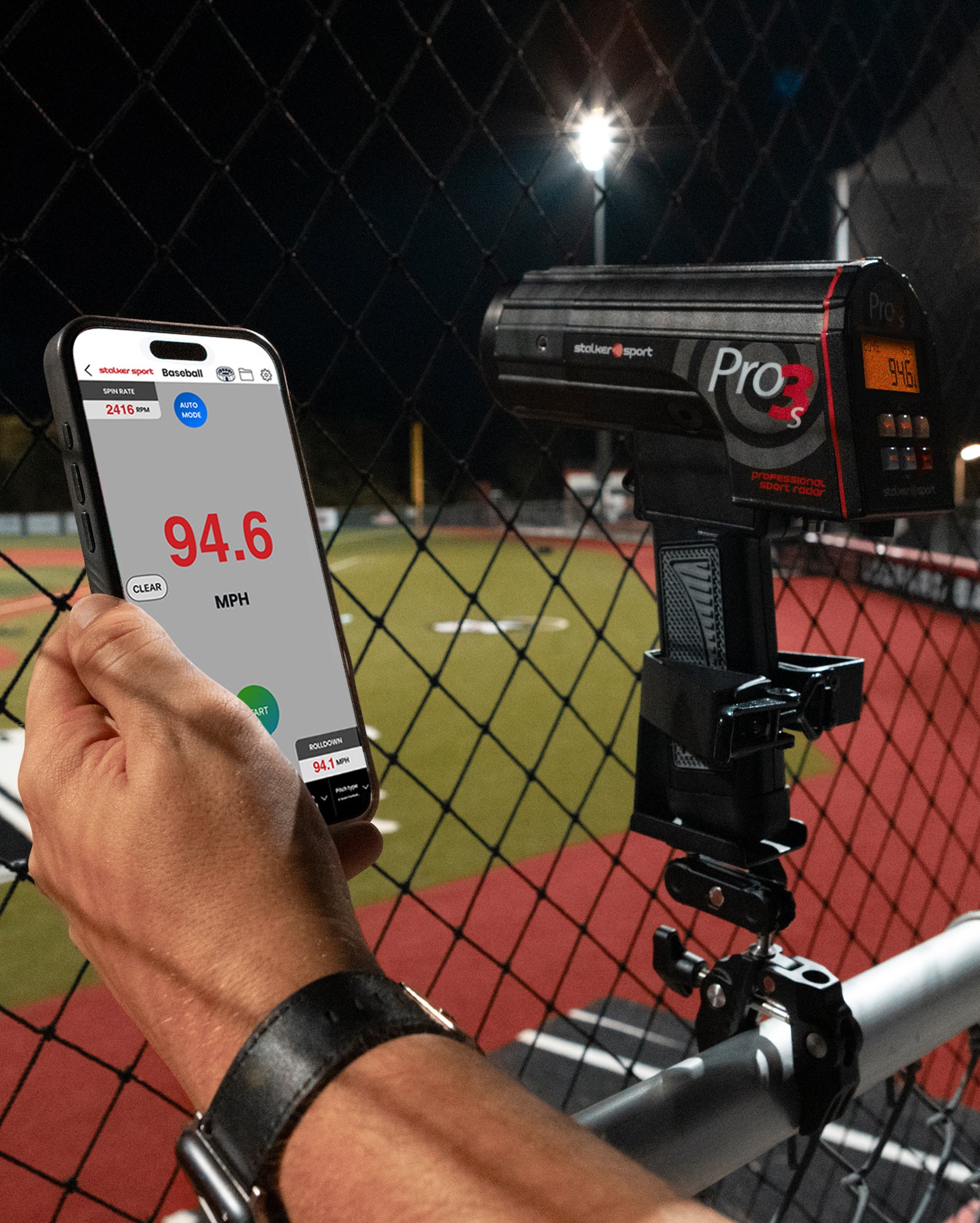 A hand holding a phone with the Stalker Sport Radar app next to a Stalker Sport Pro 3s radar gun in the 4-in-1 mount clamped to a railing at a baseball field.