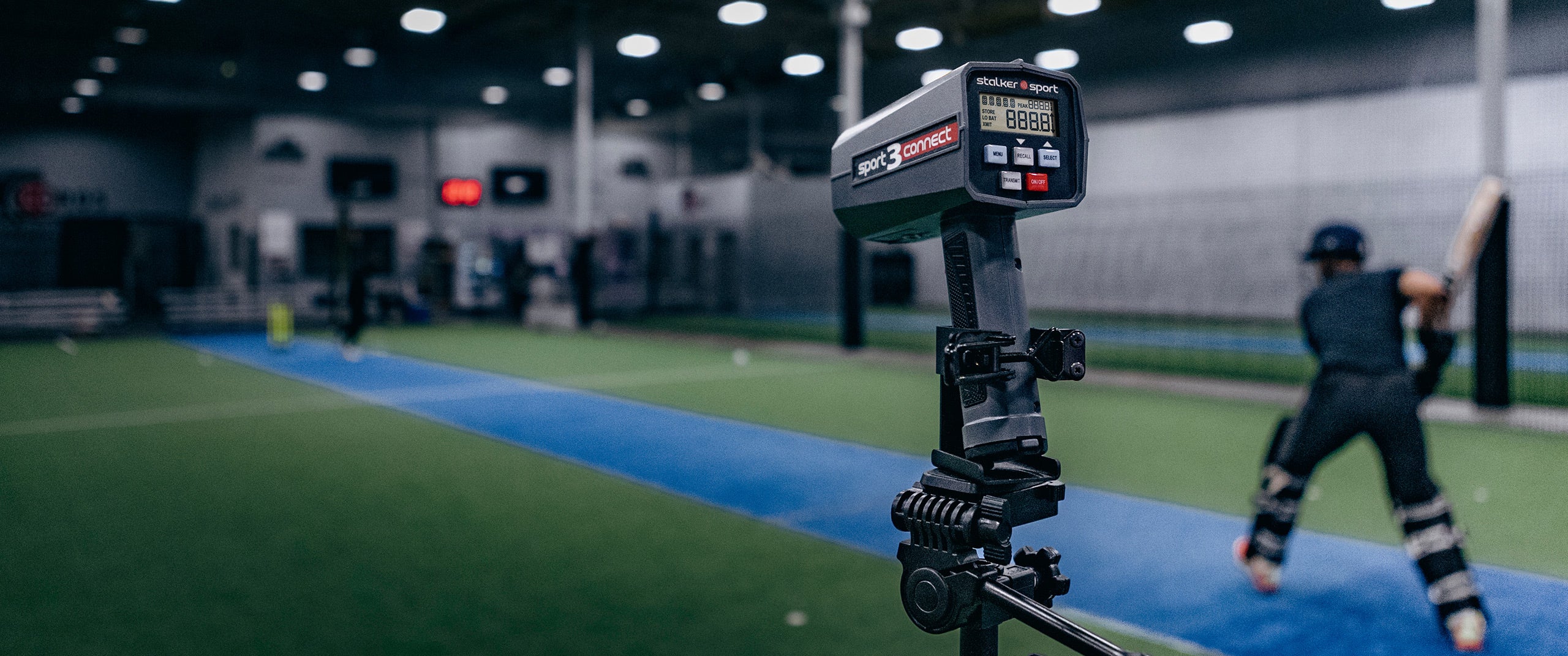 A Stalker Sport 3 Connect radar gun in a tripod measuring velocities at a cricket training facility with a cricket batter in the background.