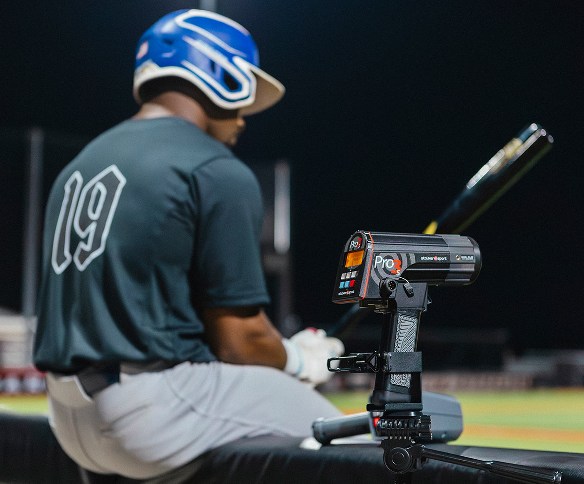 A baseball batter sitting next to a tripod-mounted Stalker Sport Pro 3s radar and a Sport 3 Connect at a baseball field at night.