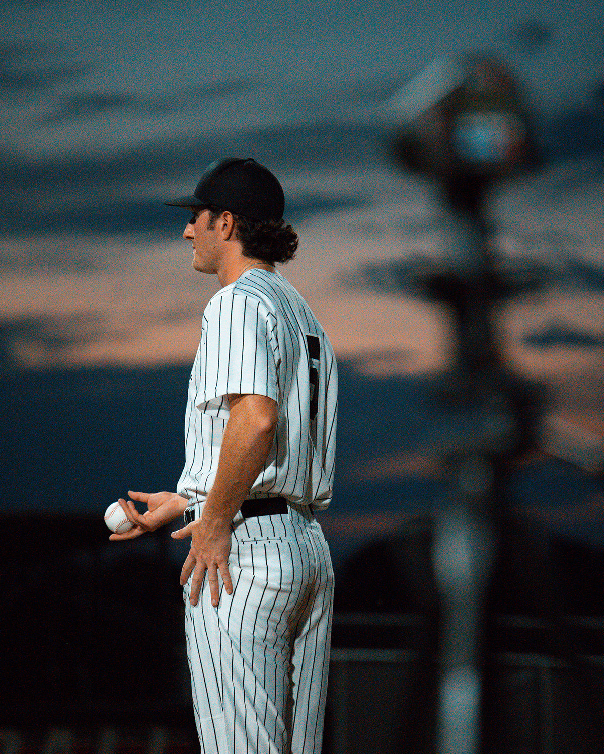 A baseball pitcher holding a baseball with an out-of-focus Pro 3s radar gun mounted on a tripod in the foreground.