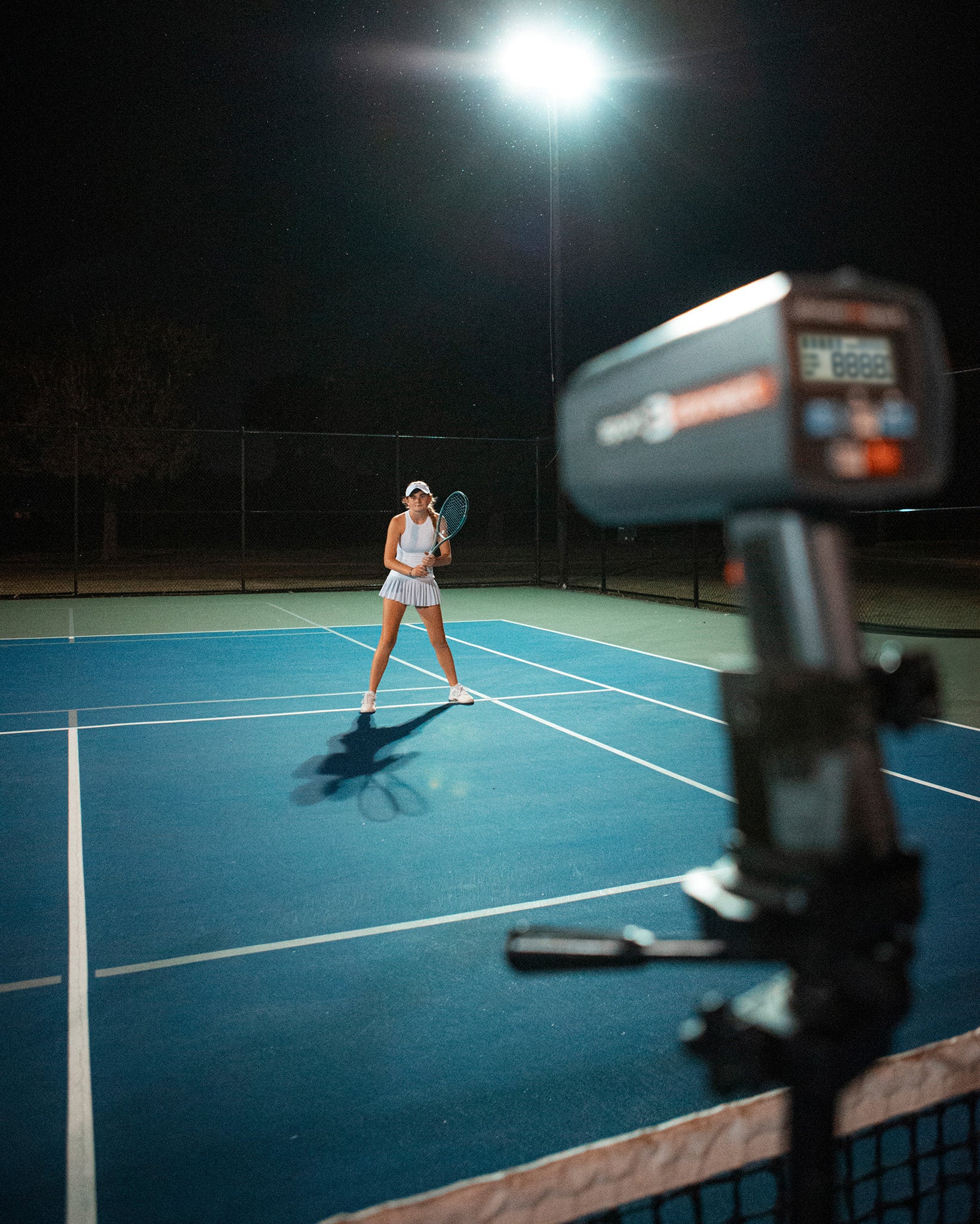 A tripod-mounted Sport 3 Connect radar gun ready to measure the return speed of a tennis player at night.
