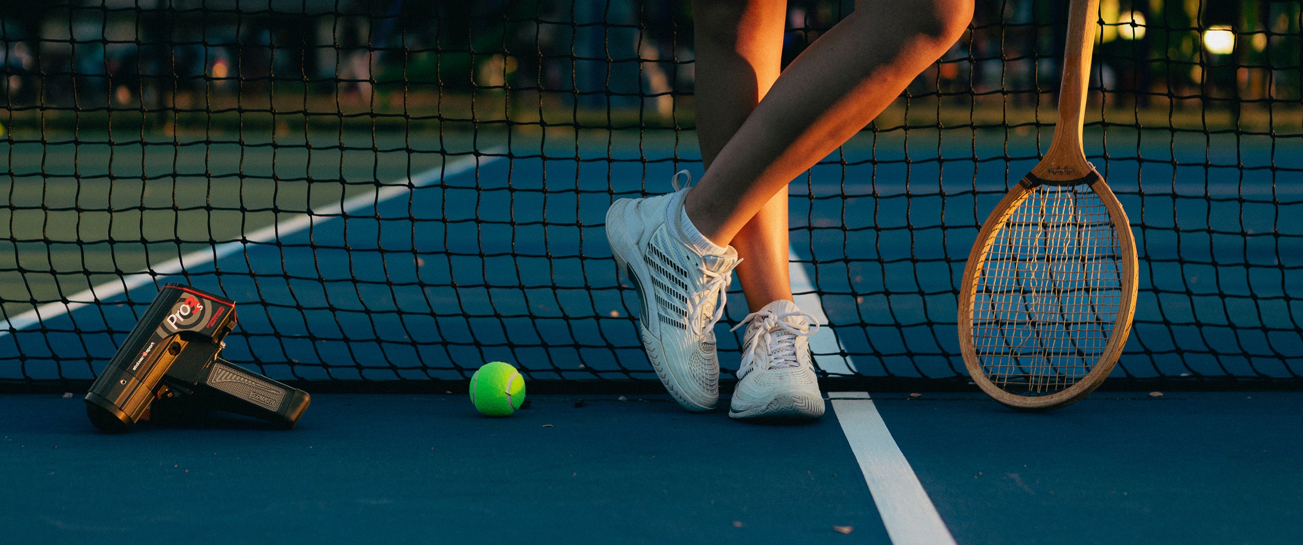 A Stalker Sport Pro 3s radar on a tennis court next to a tennis ball, tennis racket, and the feet of a tennis player. 