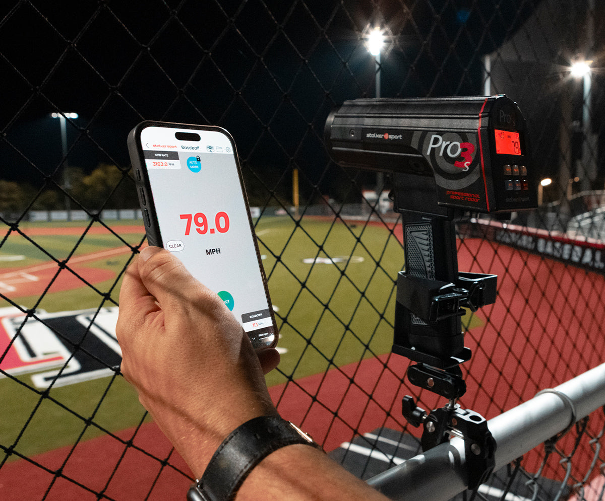 A hand holding a phone displaying the Stalker Sport Radar app next to a Pro 3s radar gun in the 4-in-1 mount clamped to a fence. Both the app and the gun are displaying a speed of 79.0 MPH. 