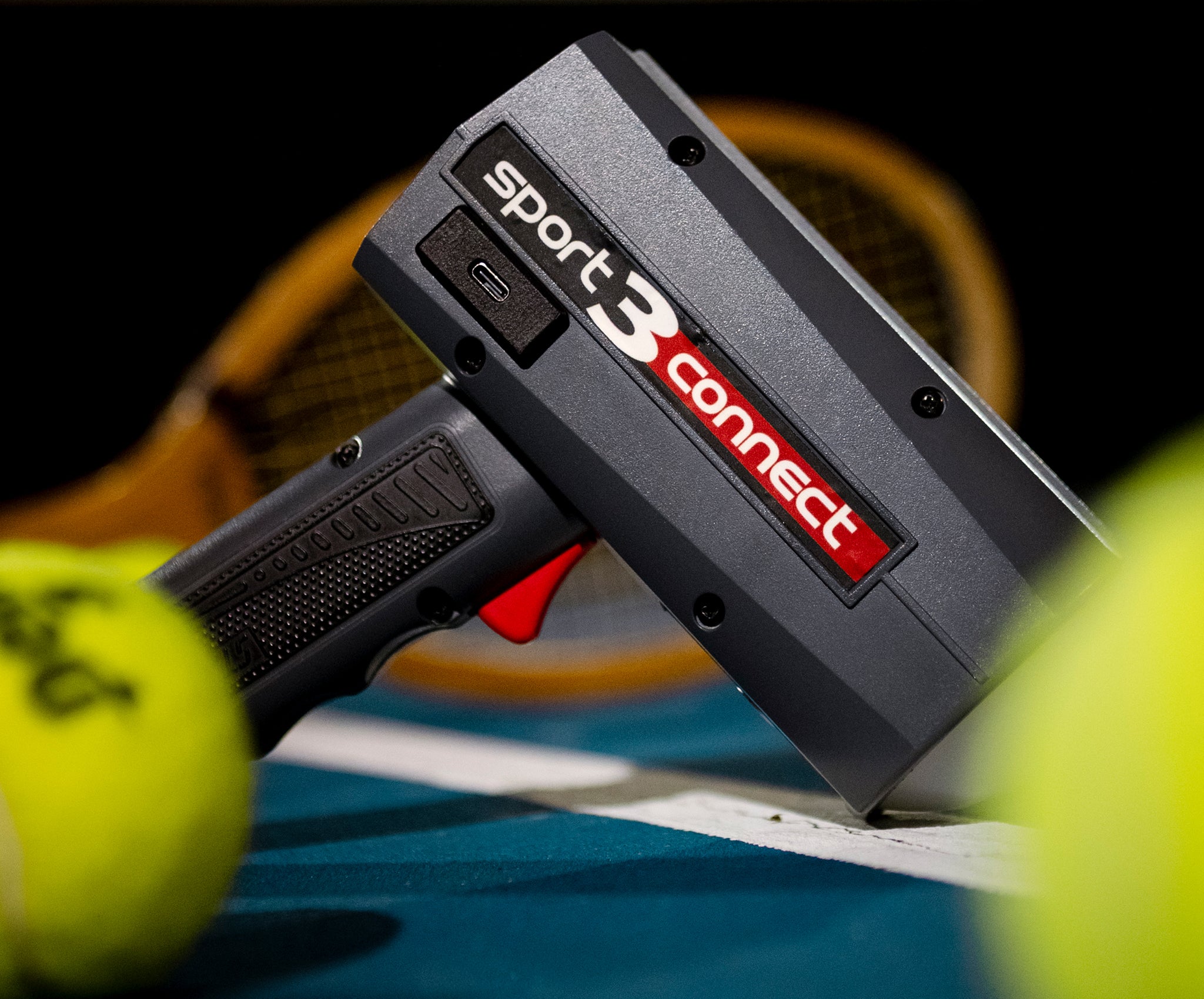 The Stalker Sport 3 Connect radar on a tennis court with tennis balls in the foreground and a tennis racket in the background. 